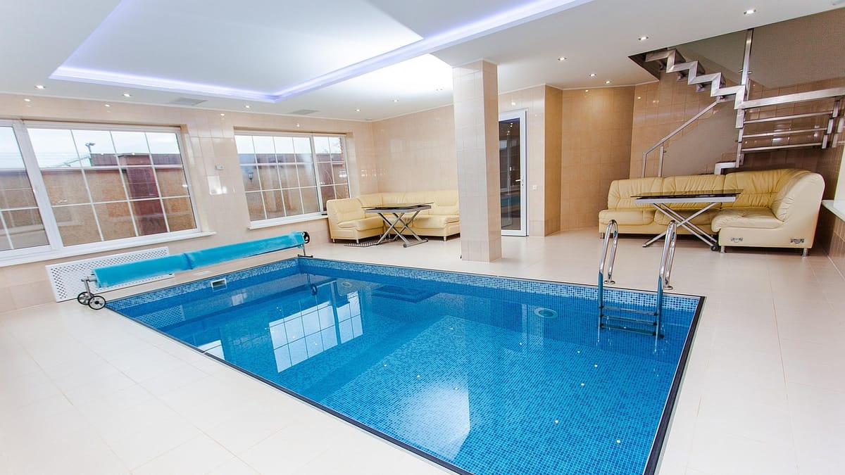 Piscine intérieure carrelée de mosaïque bleue dans une maison, avec échelle inox, enrouleur de couverture et salon de canapés beiges